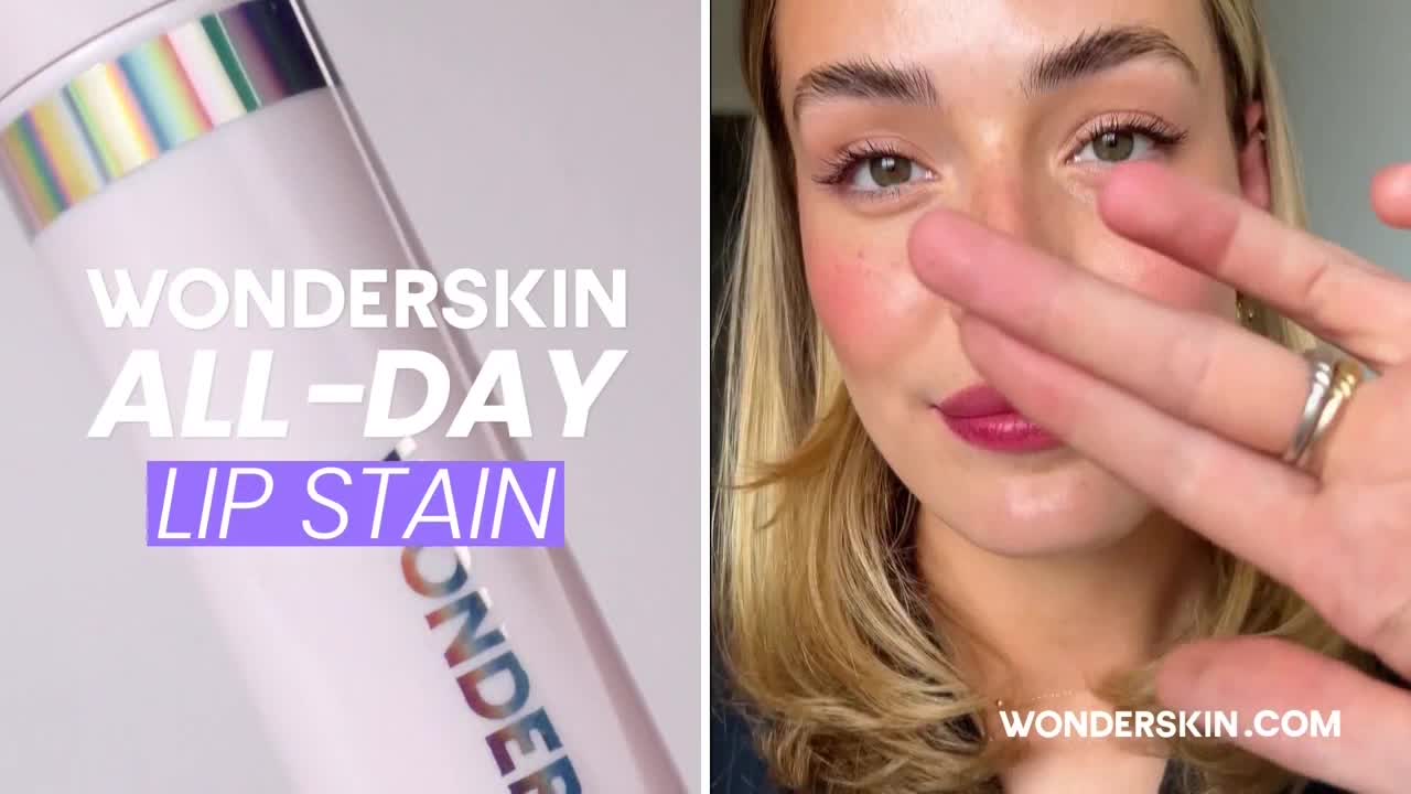 A woman's hand is held up, fingers spread, partially obscuring her face. The lip stain is a deep pink, and she's wearing a ring.
