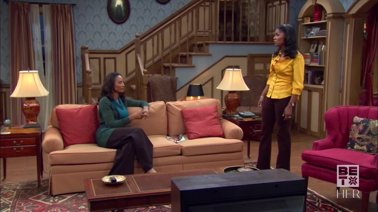 A woman in a yellow shirt stands near a red chair, facing another woman seated on a sofa. The woman on the couch, wearing a green sweater, looks towards her.
