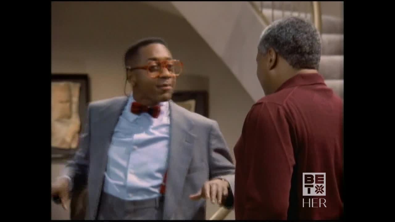 A man in a gray suit and bow tie gestures with his hands as he speaks to another man. The other man, in a maroon shirt, listens intently near a staircase.
