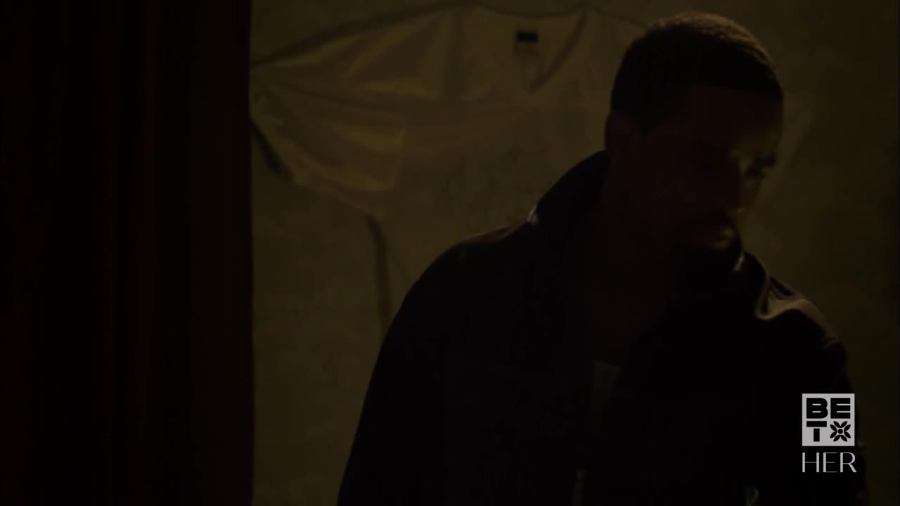 A man in a dark jacket is looking down, his face mostly obscured by shadow. He's standing near a wall, and the scene is dimly lit.
