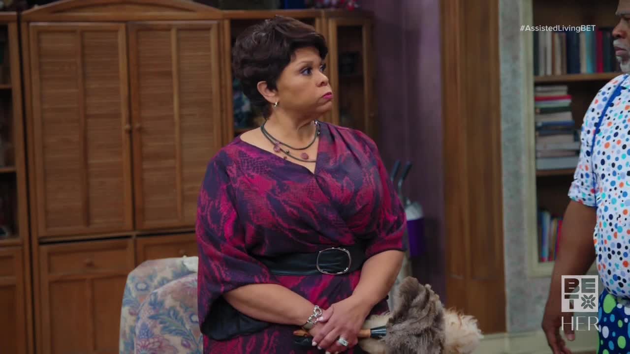 A woman in a patterned dress stands with her arms crossed, her gaze fixed off-camera. Beside her, a man in a polka-dot shirt is partially visible, both seemingly in a scene from BET Her's *Assisted Living*.
