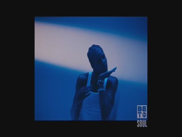 The man in the white tank top holds a microphone, his hand near his mouth. A blue and white backdrop provides the stage for this BET Soul performance.
