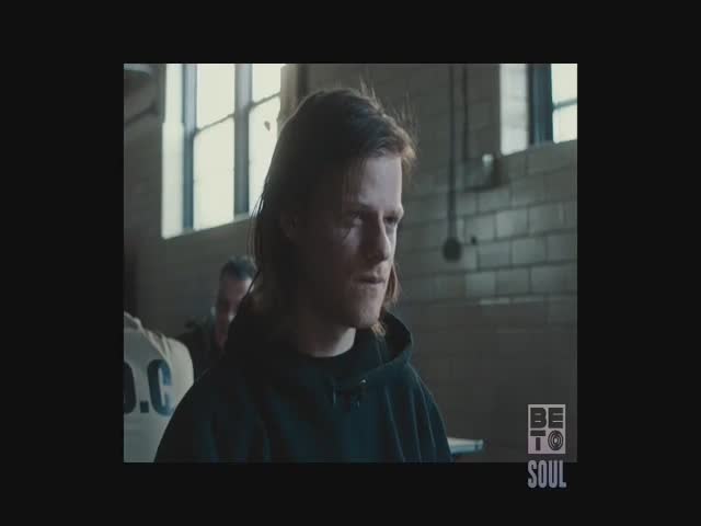 The man in the black hoodie looks toward the camera, his expression neutral. Behind him, a person in a white shirt with "D.C." printed on it stands in the background.
