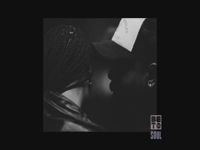Two people are close, their faces nearly touching. One wears a cap with "LOS ANGELES" visible, the other has their hair in tight braids.
