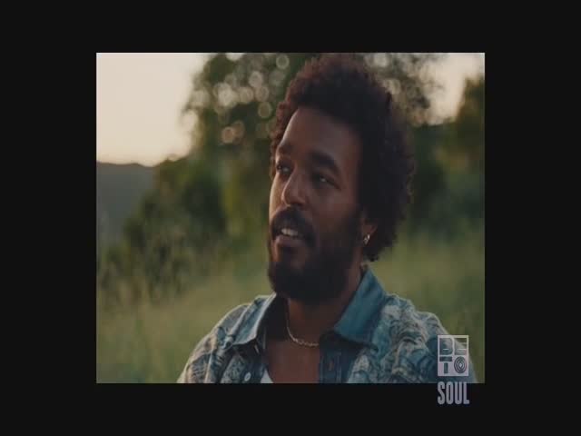A man with dark, curly hair sits in a field, looking off-camera with a slight smile. The BET Soul logo is visible in the corner, suggesting a music video or program from the United States.
