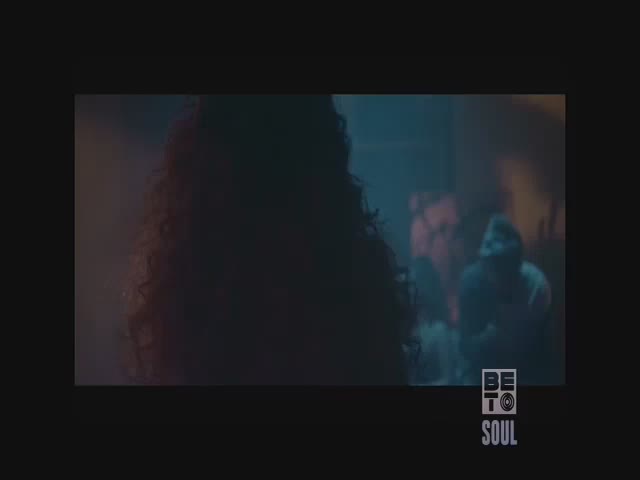 A person with curly hair is turned away from the camera, their silhouette dominating the frame. In the background, a figure in a hoodie stands in a dimly lit space, possibly on a BET Soul music video set.

