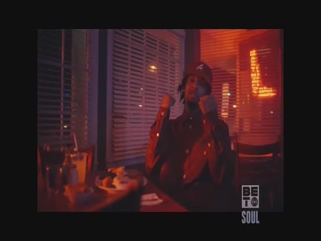 A man in a maroon cap sits at a table, his fists clenched. The scene has a warm, reddish glow, like it's a late-night spot on BET Soul.
