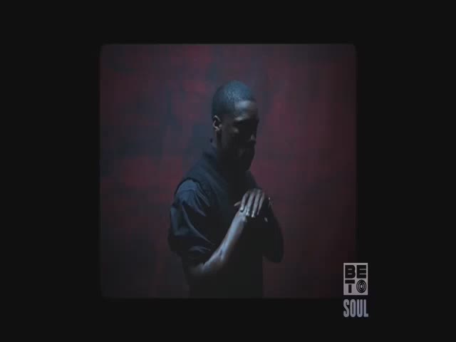 A man stands in a dim space, his hands clasped in front of him. Behind him, a hazy red and black backdrop provides the only light.

