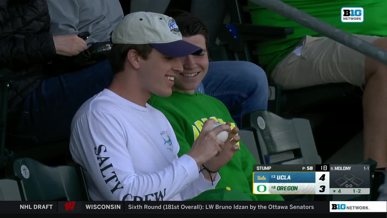 Two guys in the stands are smiling, one holding a baseball. The Big Ten Network score shows UCLA leading Oregon.
