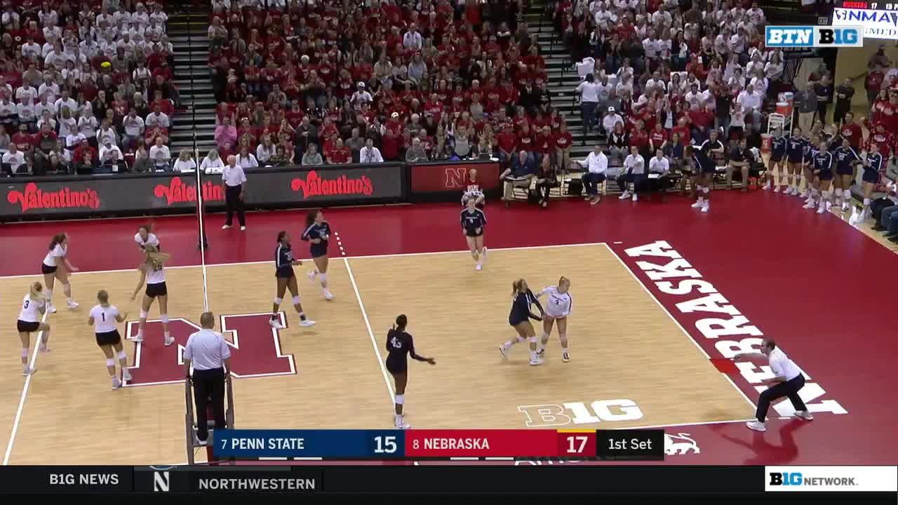 A player in white prepares to hit the volleyball over the net, as a teammate in black gets ready to assist. The Nebraska team, in red and white, watches closely.
