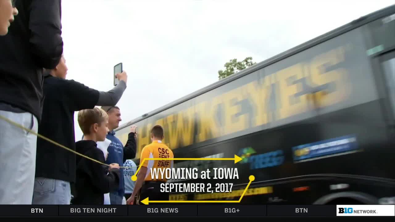 A team bus with "HAWKEYES" in yellow lettering speeds past a small crowd. A man in a yellow staff shirt stands near the bus, while others hold up phones.
