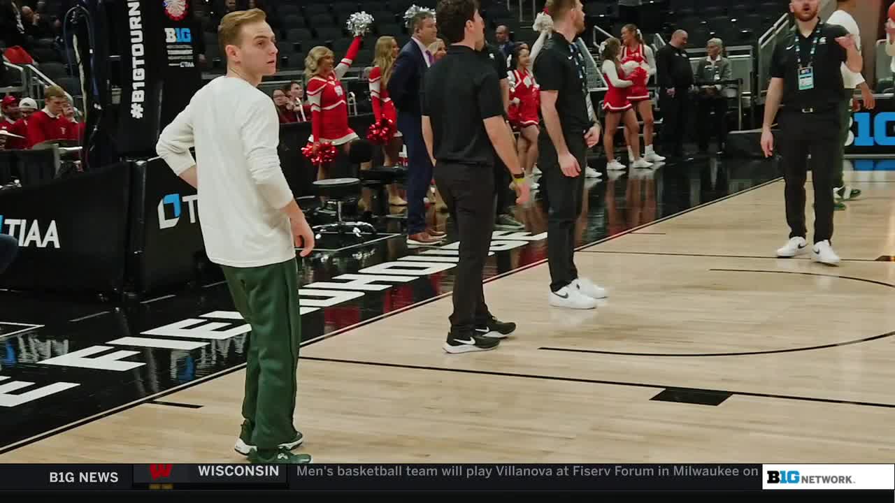 A man in green sweats stands near the court, his hands on his hips. Other staff members in black shirts and pants stand nearby, while cheerleaders in red and white wait in the background.
