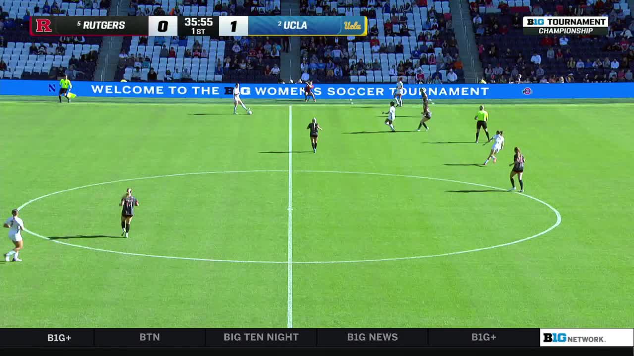 A UCLA player in white is dribbling the ball toward the center of the field, while a Rutgers player in black and red chases her. The score is 1-0 in the first half of the Big Ten Women's Soccer Tournament.
