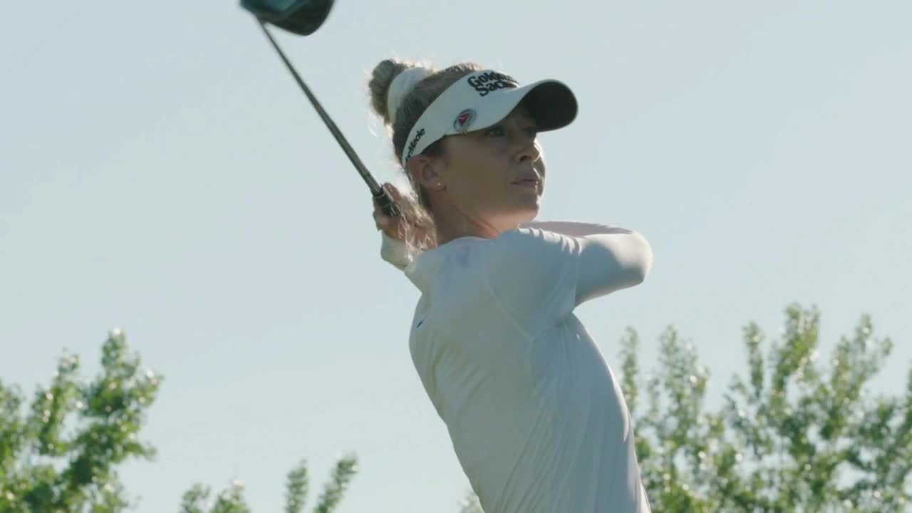 The golfer is mid-swing, the club head arcing upward against the bright sky. Her focus is intense, and the movement looks powerful.
