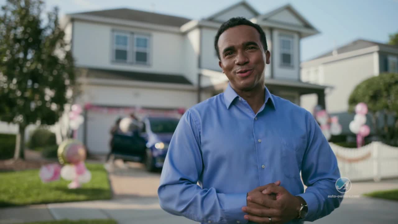A man in a blue shirt smiles at the camera in front of a house decorated with pink balloons. A dark blue car sits in the driveway, and people are getting out of it.

