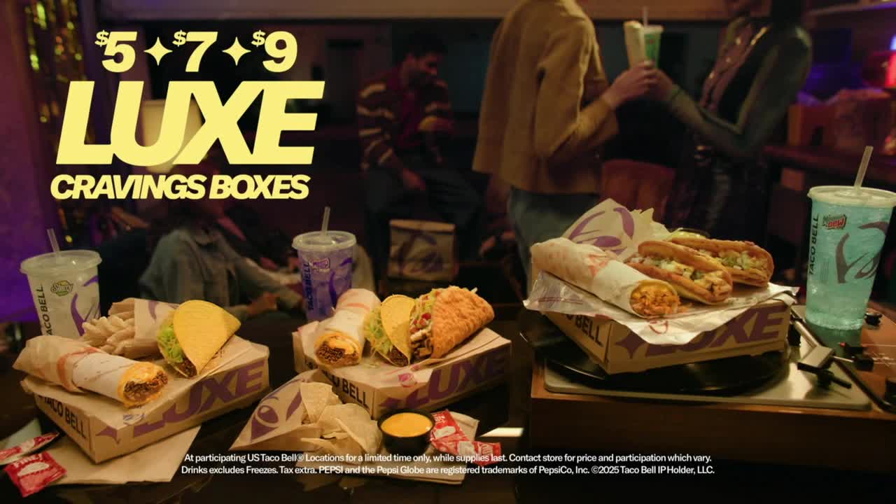 Taco Bell's Luxe Cravings Boxes are displayed with various tacos and burritos, alongside drinks, on a table with people in the background. The advertisement highlights the different box options and prices.
