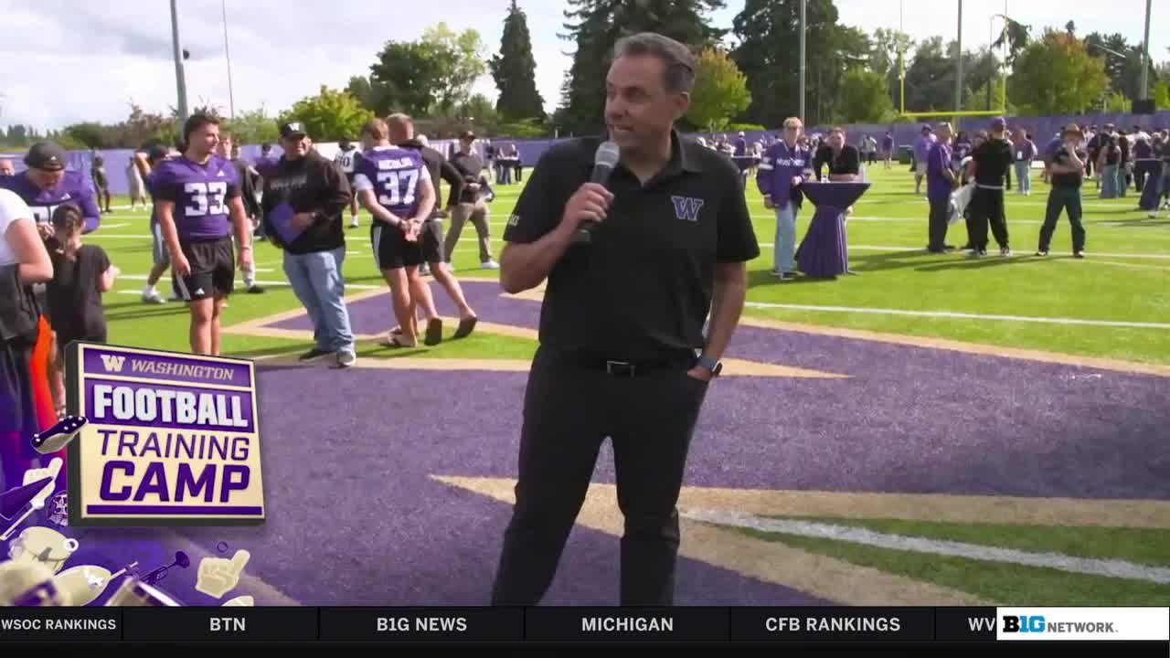 A man in a black polo shirt with a microphone stands on the field, addressing the camera. Behind him, a group of football players in purple jerseys are milling around on the green grass.
