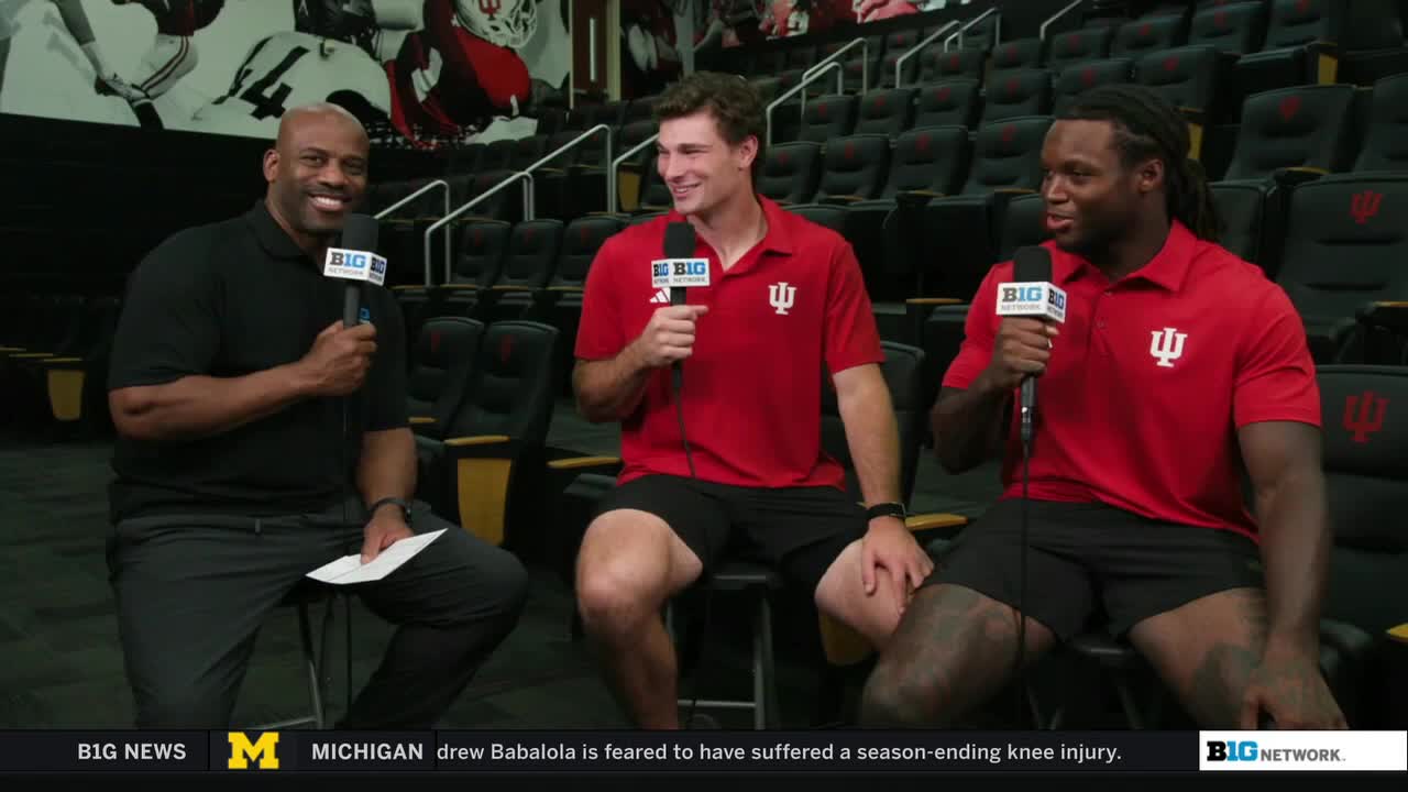 Three men sit in a row, each holding a microphone with the Big Ten Network logo. The two on the right wear matching red shirts with the Indiana University logo.
