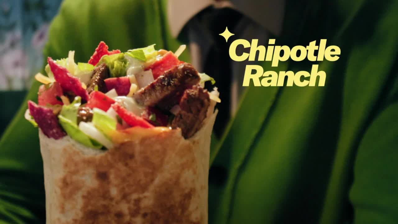 A tightly-wrapped burrito, overflowing with colorful fillings, is held up in front of a person wearing a green jacket. The words "Chipotle Ranch" are displayed with a starburst effect.
