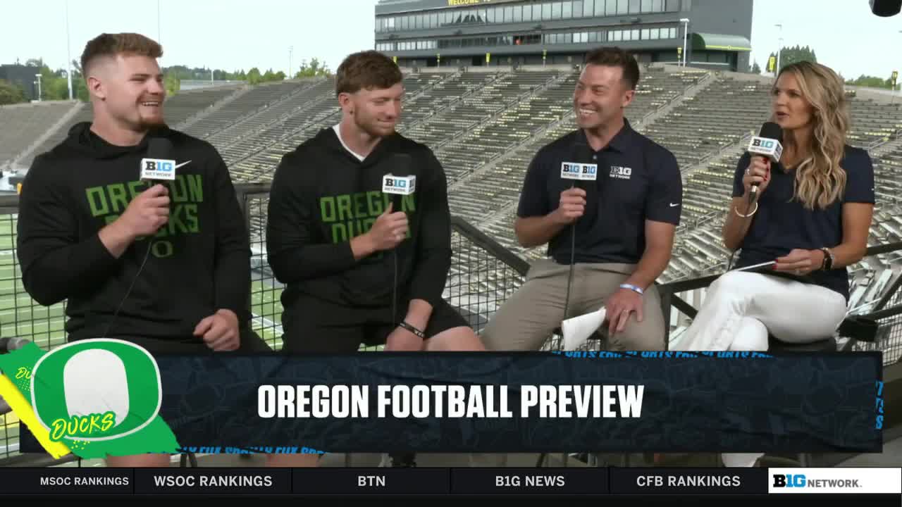 Four people sit on a platform, each holding a microphone with the Big Ten Network logo. The two men on the left wear Oregon Ducks hoodies, and the woman on the right holds a paper, presumably a script, as she speaks.
