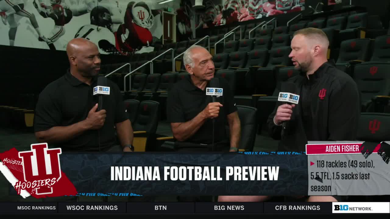 Three men sit in a row, each holding a microphone with a "Big Ten" logo. A man in a black jacket is speaking, while the other two listen intently.
