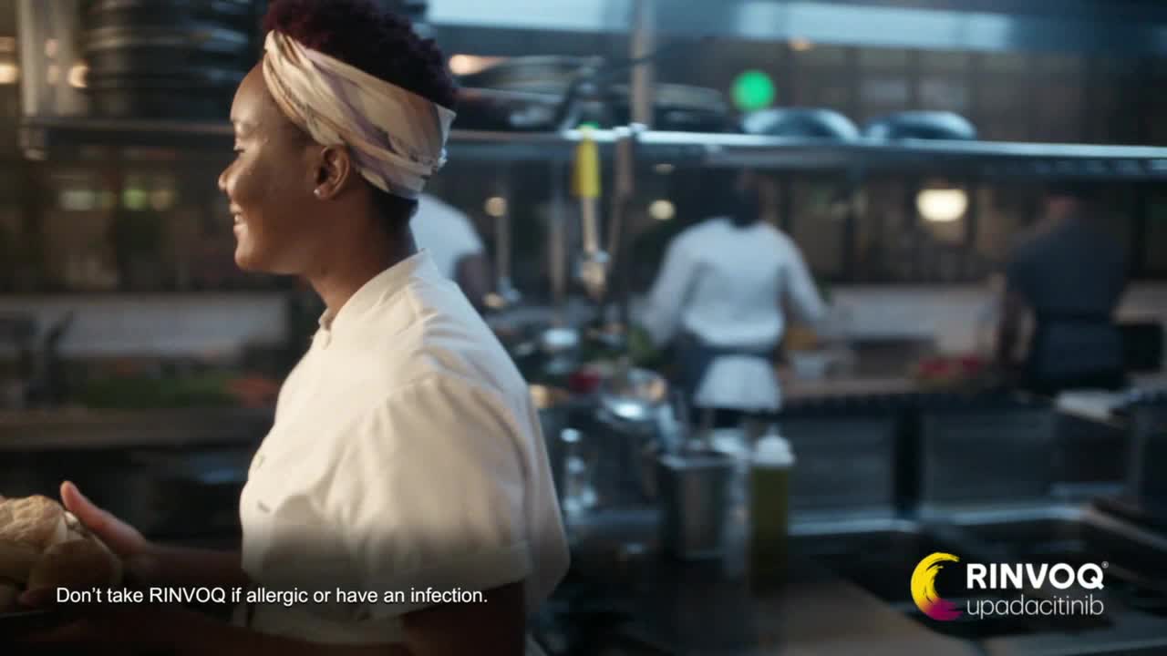 A chef in a white coat smiles, holding a basket of bread. Other cooks move in the background of a commercial kitchen.
