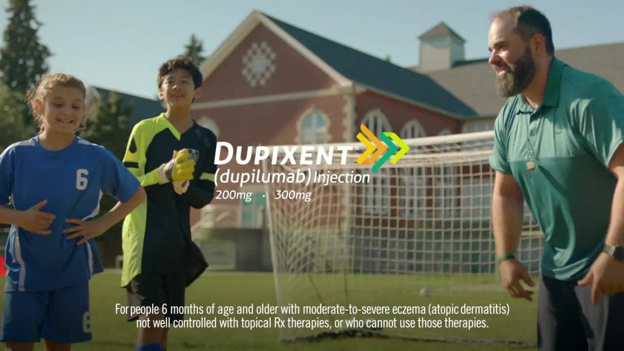 A man in a green shirt grins as he looks at two kids, a girl in blue and a boy in a goalie uniform. The three of them are on a soccer field, with a goal and a school building in the background.

