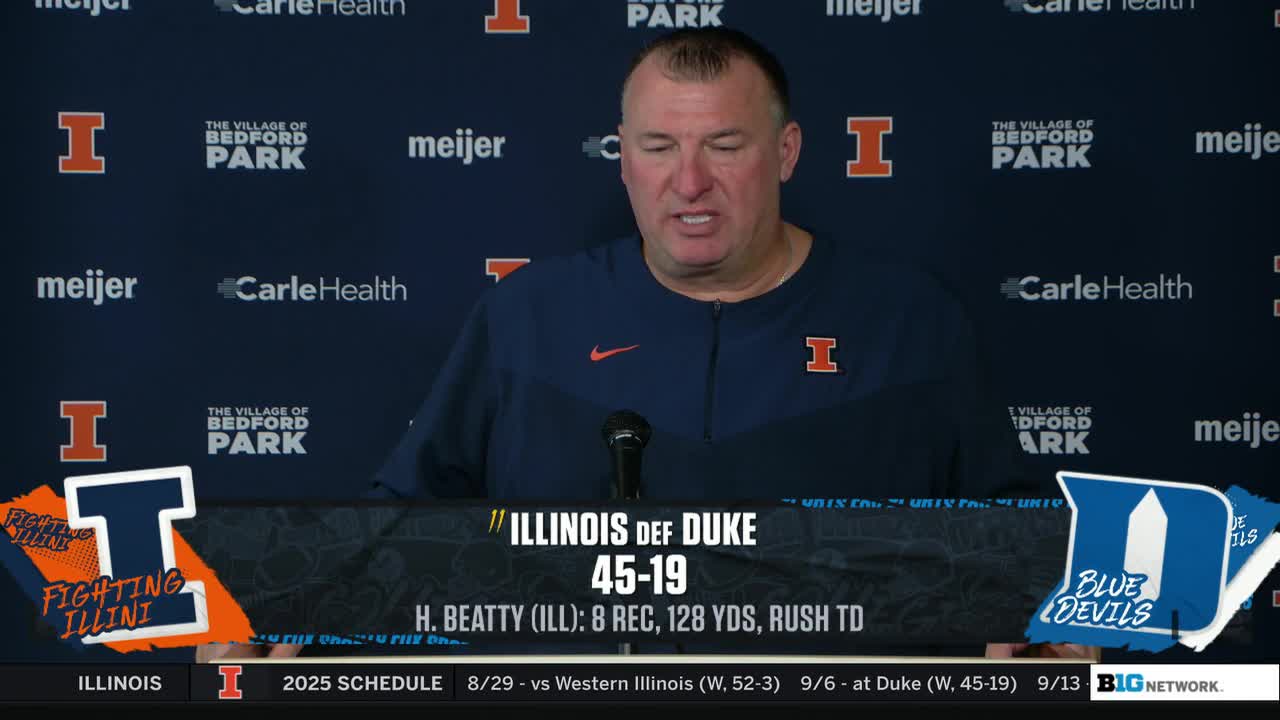 The Illinois coach is speaking into a microphone, his face lit by studio lights. A graphic on the Big Ten Network screen shows the final score of the Illinois-Duke game.
