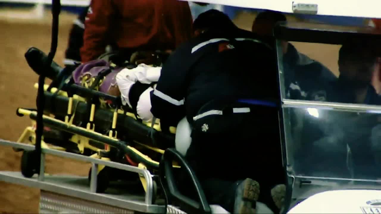 A person lies on a stretcher, covered in a blanket, as medical personnel attend to them. The stretcher is being loaded onto a cart, probably for transport.
