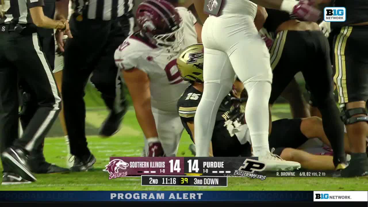 A pile of players, mostly legs and helmets, has formed near the sideline. A player in a black and gold uniform is on the ground, while others stand over him.
