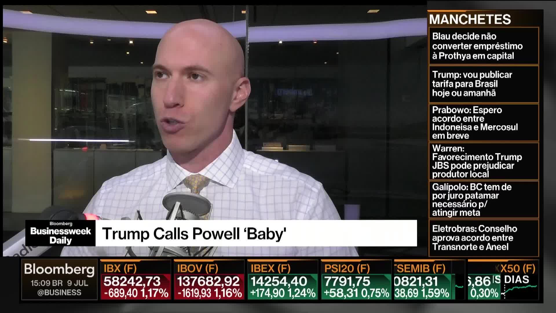 On Bloomberg TV in the United States, a man in a button-down shirt is speaking, his mouth open, and a microphone is positioned in front of him. The lower third of the screen displays the headline "Trump Calls Powell 'Baby.'"
