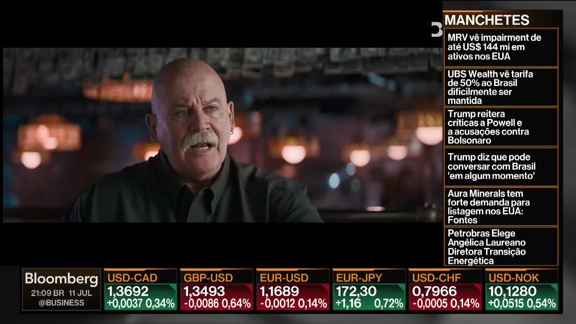 A man with a white mustache is speaking, his mouth open, gesturing with his hands. The Bloomberg TV screen displays currency rates and news headlines in Portuguese.
