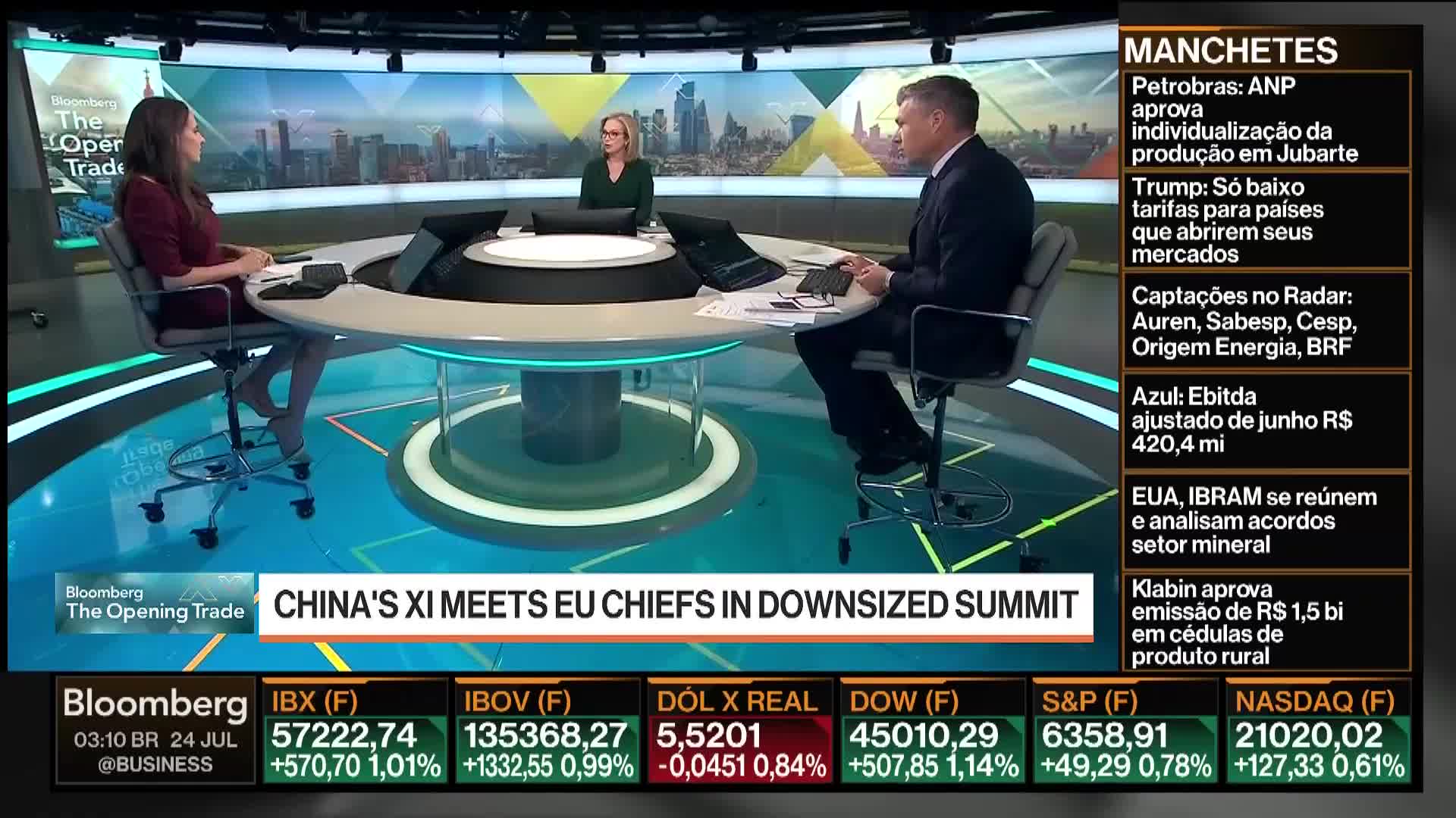 Three people sit around a curved desk, discussing the news on Bloomberg TV. The man in the suit gestures with his hands as he speaks, while the woman to his left listens intently.
