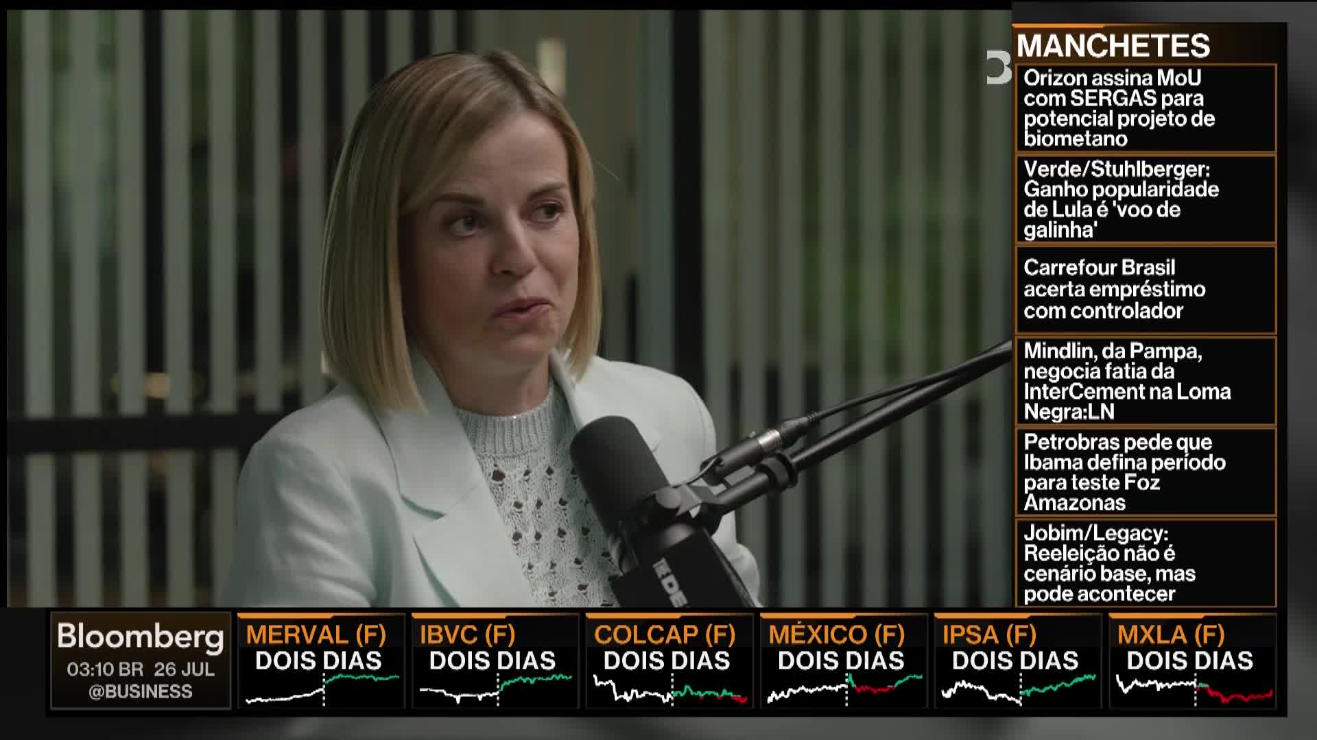 A woman in a light blue blazer speaks into a microphone on a Bloomberg TV set. The screen displays financial news headlines and stock market data from various Latin American exchanges.
