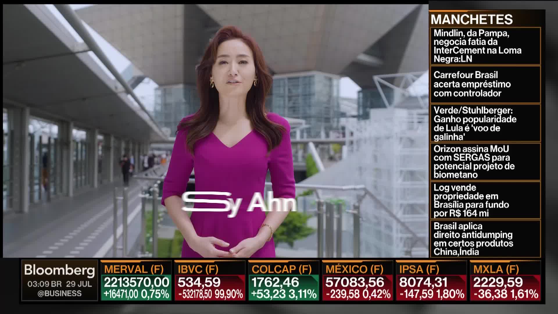Sy Ahn, in a magenta dress, stands in front of a modern building, speaking directly to the camera. The Bloomberg logo and stock market data are displayed at the bottom of the screen.
