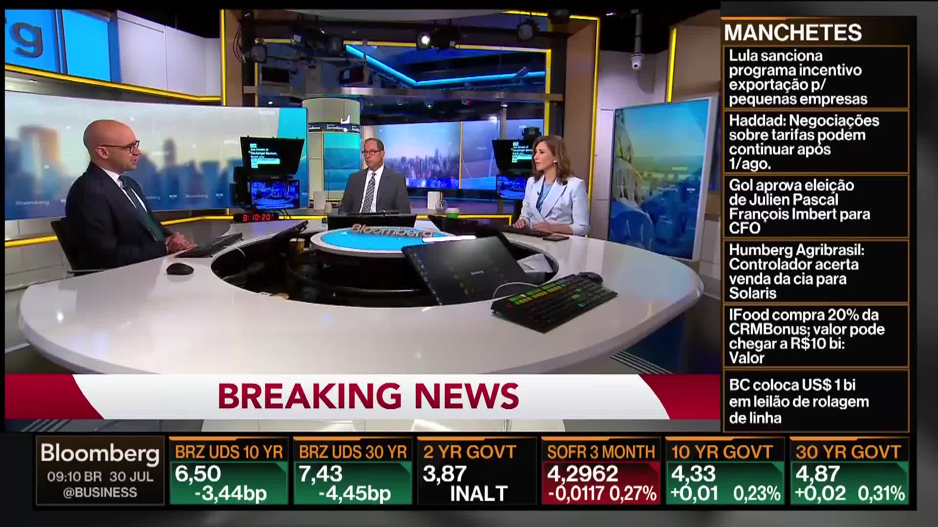 The Bloomberg news set features three people at a curved desk, with a "Breaking News" banner displayed. The broadcast focuses on financial news, with headlines and market data scrolling on the screen.
