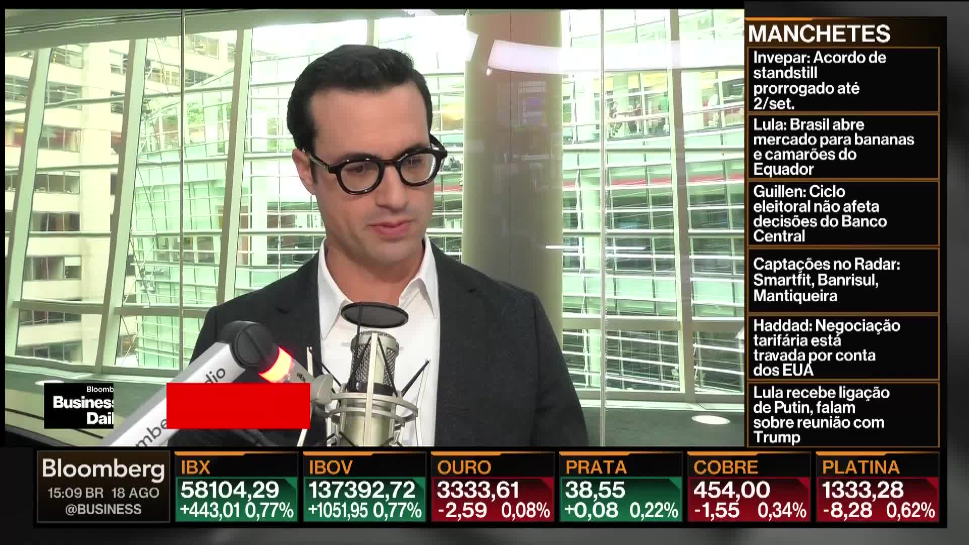 A man in glasses speaks into a microphone, framed by the Bloomberg Business Daily logo and financial data tickers. News headlines scroll on the right side of the screen.
