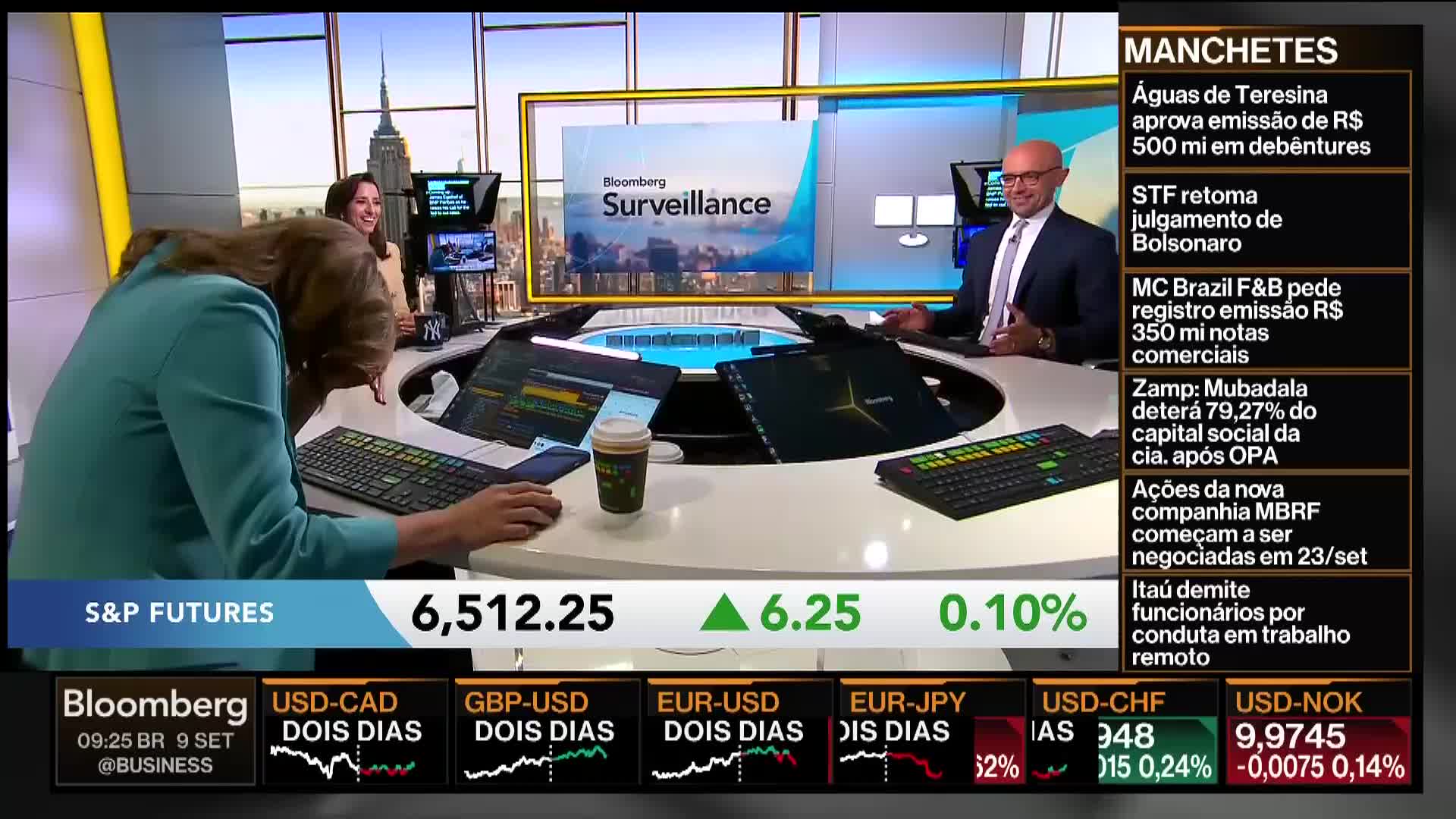A woman in a teal blazer hunches over a keyboard, focused on the screen in front of her. Across the set, a man in a suit gestures as he speaks, likely on Bloomberg TV from the United States.
