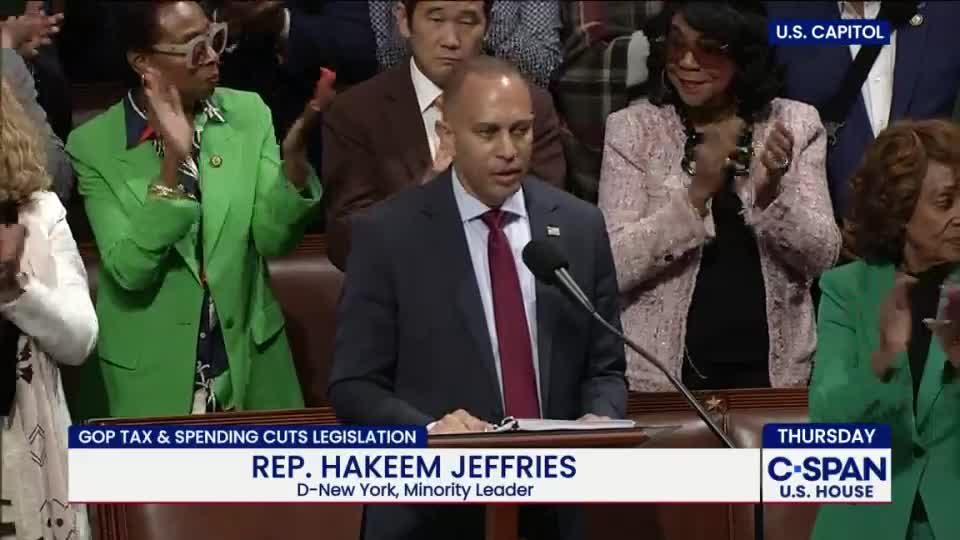 Rep. Hakeem Jeffries, in a dark suit and red tie, speaks into a microphone at a podium. Applause erupts around him from several people in the background, as seen on C-SPAN.
