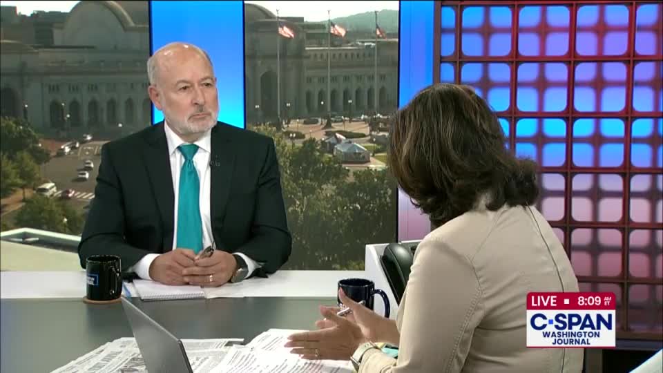 A man in a suit, with a bright teal tie, listens intently as a woman gestures with her hands. The C-SPAN Washington Journal logo is visible at the bottom right, and the backdrop shows a building with American flags.
