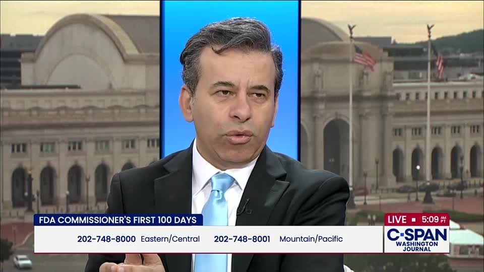 A man in a suit speaks directly to the camera, likely on a C-SPAN broadcast from Washington, D.C. Behind him, a backdrop shows a government building and American flags.
