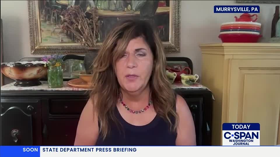 A woman in a dark top speaks directly into a camera, likely for C-SPAN, with a backdrop of a decorated shelf and a framed painting. The lower-third graphic indicates a State Department press briefing is coming up.
