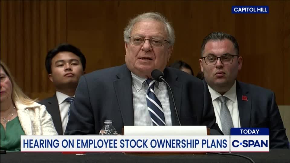 A man in a suit and tie speaks into a microphone at a hearing on Capitol Hill, as broadcast by C-SPAN. Two other men in suits flank him, listening intently.

