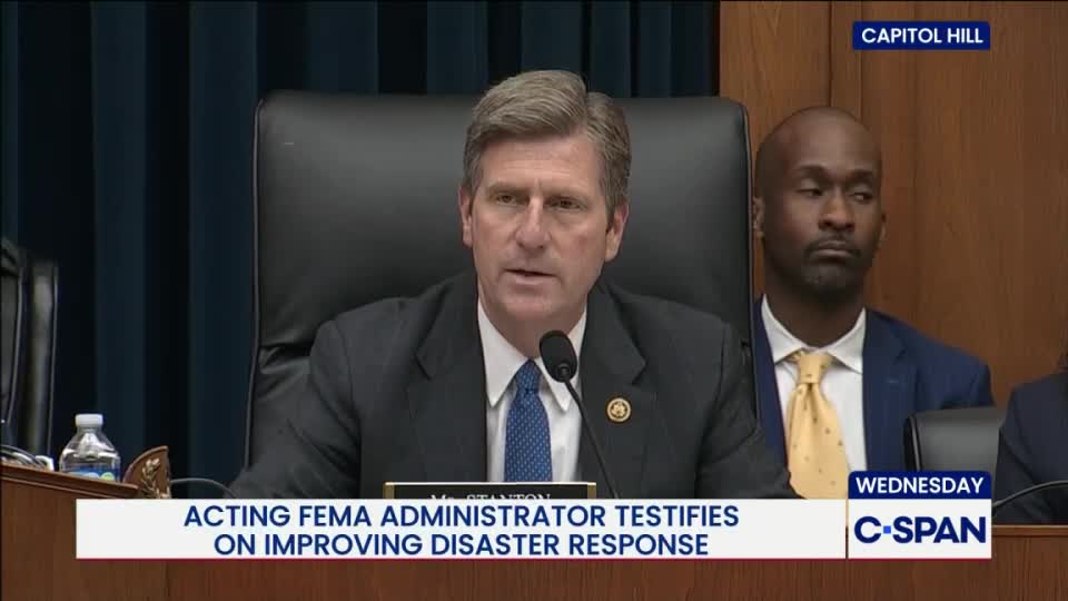 The Acting FEMA Administrator, speaking into a microphone, is in the middle of a hearing on Capitol Hill, broadcast by C-SPAN. A man in a blue suit and yellow tie sits to his right, listening intently.
