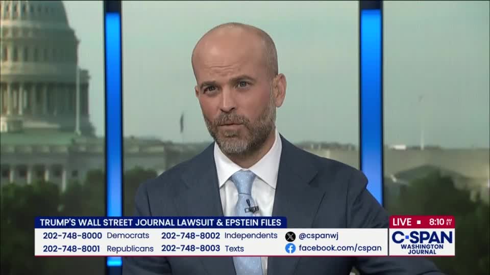 A man in a suit and tie looks directly at the camera, speaking on C-SPAN. Behind him, a window reveals a view of a building in Washington, D.C.
