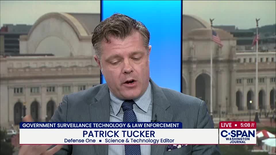 Patrick Tucker, a Science & Technology Editor, is speaking on C-SPAN about government surveillance. Behind him, a backdrop shows a building with American flags waving.
