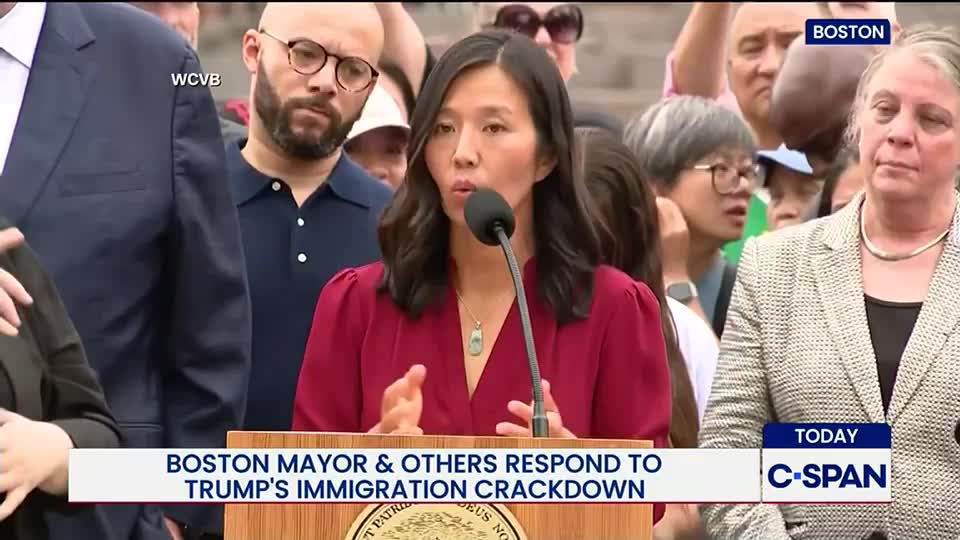 The Boston mayor, wearing a red blouse, speaks into a microphone, gesturing with her hands. A crowd of people, some looking on intently, surrounds her as C-SPAN broadcasts the event.
