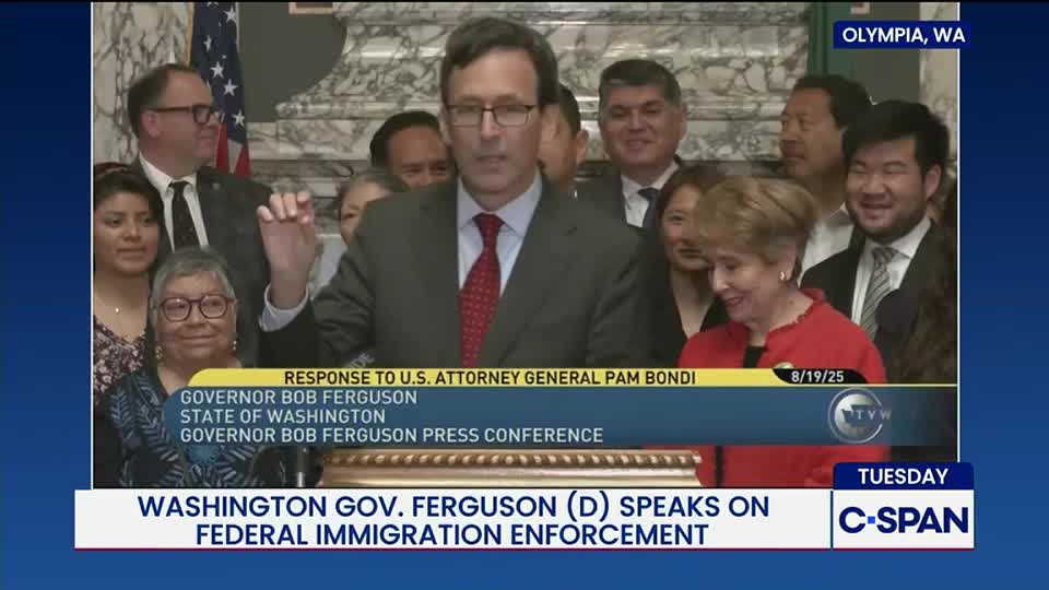 Governor Bob Ferguson of Washington, speaking on federal immigration enforcement, gestures with his right hand. A crowd of people, some smiling, stand behind him in a room, captured by C-SPAN.
