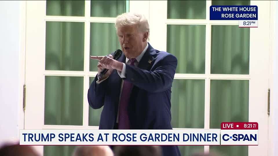 He's holding a microphone, gesturing emphatically with his right hand. The C-SPAN feed shows him speaking at a Rose Garden dinner in the United States.
