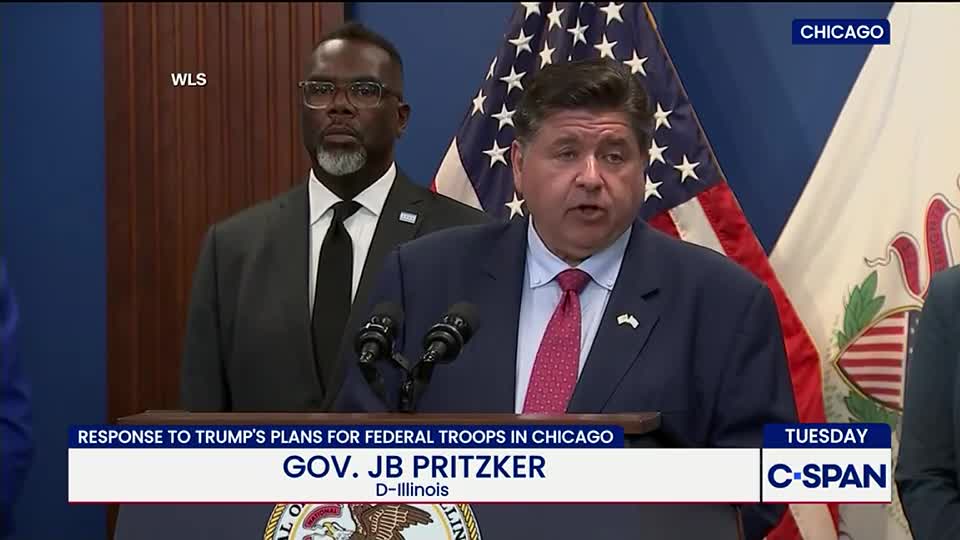 Governor Pritzker, wearing a blue suit and red tie, speaks at a podium in front of an American flag. A man in a dark suit stands behind him, watching the press conference, which is being broadcast on C-SPAN.

